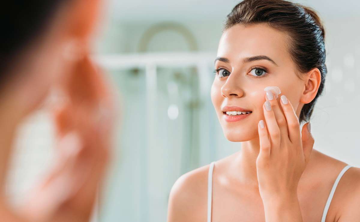 Donna che applica la crema idratante sul viso davanti allo specchio per preparare la pelle alla base trucco