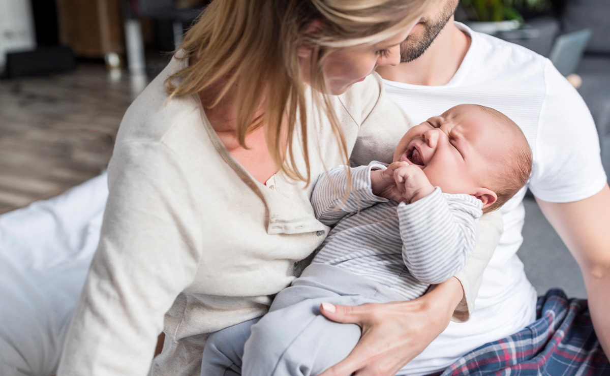 Neonato che piange in braccio a due genitori preoccupati seduti insieme, scena di comfort e accudimento in un ambiente domestico.