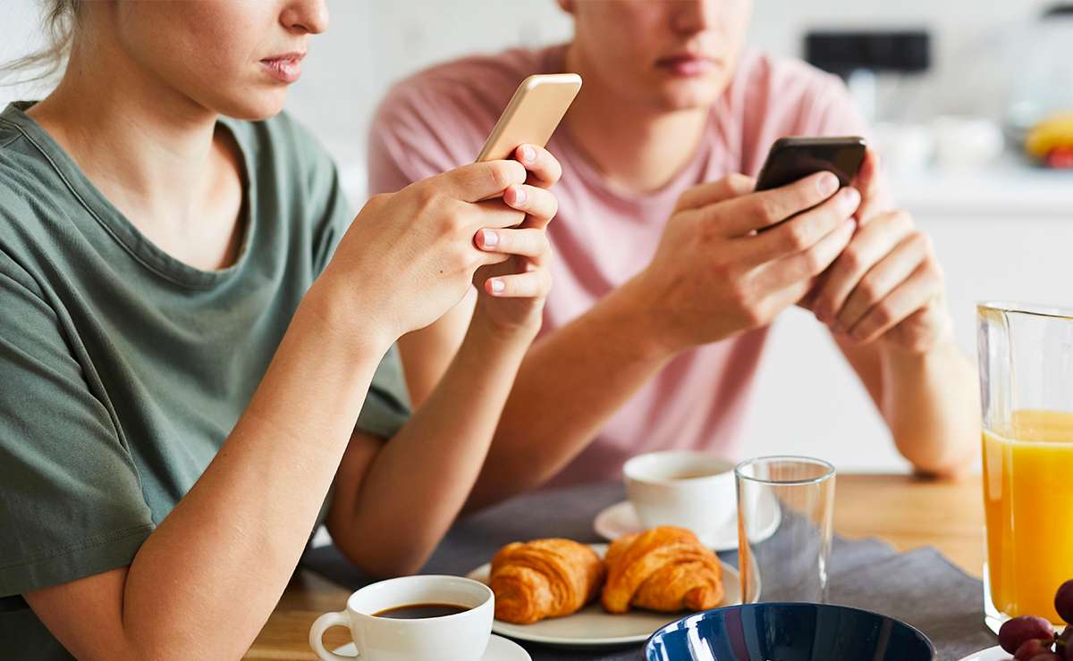 Un uomo e una donna seduti al tavolo della colazione con lo sguardo perso ognuno sullo schermo del proprio telefono