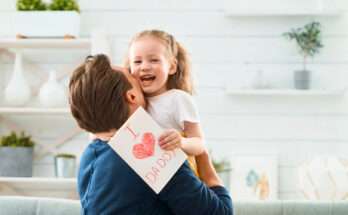 Padre abbraccia la figlia sorridente mentre lei tiene un biglietto con la scritta “I love Daddy”, simbolo d’affetto per la Festa del Papà.