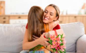 Madre e figlia si abbracciano sorridenti sul divano durante la Festa della Mamma. La mamma riceve un regalo con un fiocco rosso e un mazzo di fiori rosa.