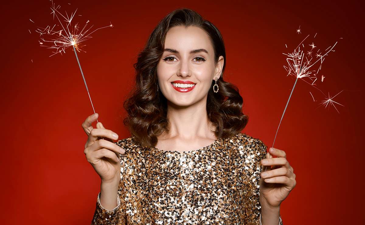 Donna sorridente in abito scintillante dorato tiene due stelline luminose accese, su sfondo rosso, simbolo di festa e buon auspicio per il Capodanno.