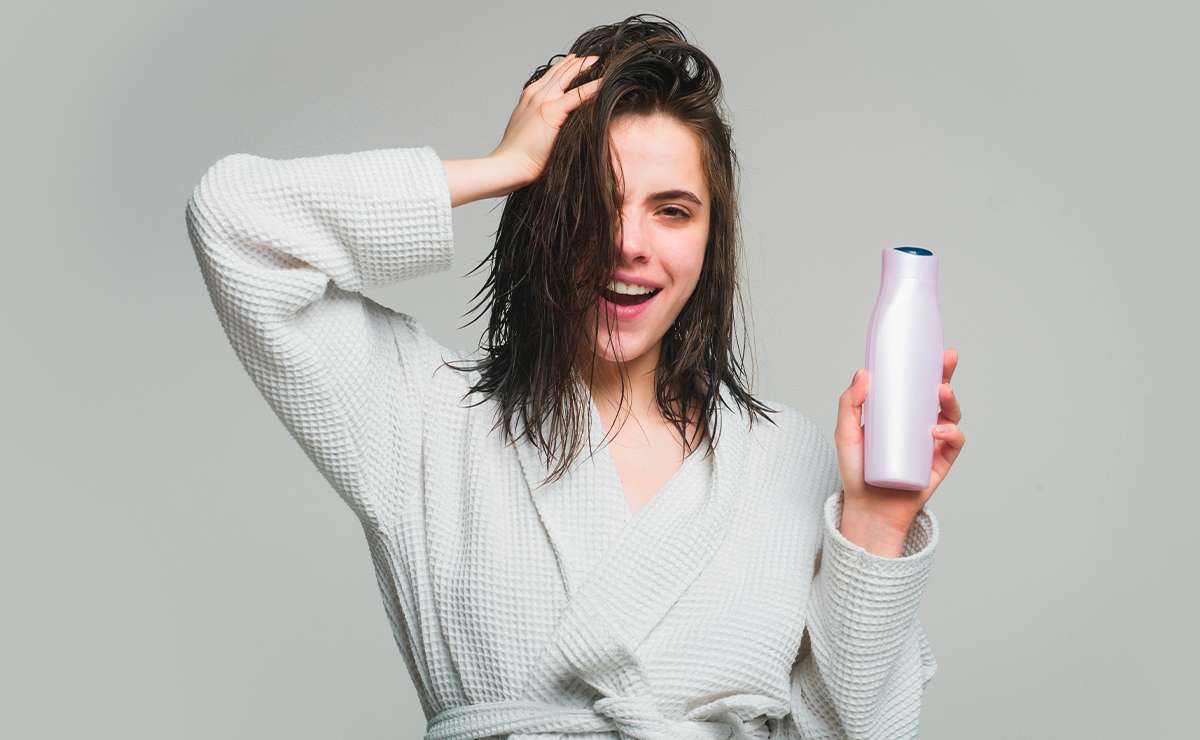 Donna con capelli bagnati e accappatoio bianco sorride mostrando uno shampoo: come scegliere lo shampoo giusto per i propri capelli.