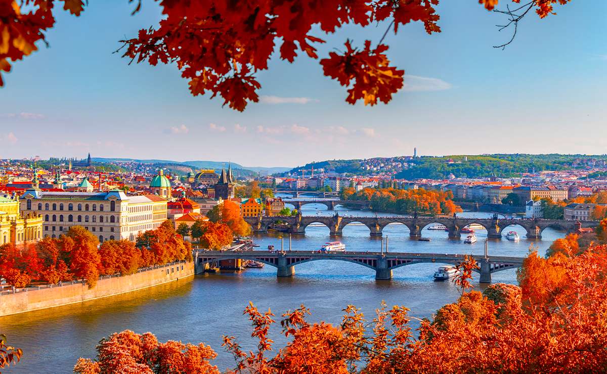 Veduta panoramica di Praga in autunno con foglie rosse in primo piano, il Ponte Carlo e i tetti storici illuminati dalla luce calda della stagione.