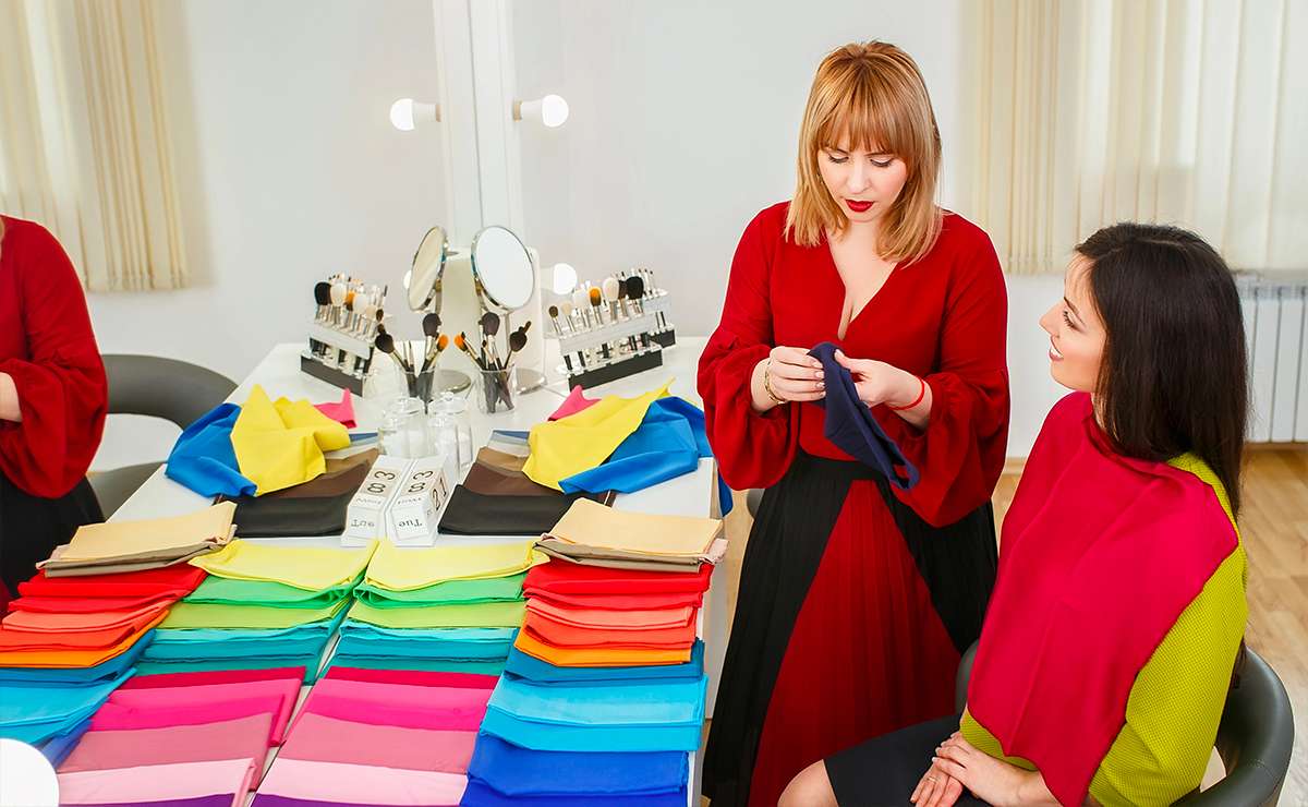 Consulente di armocromia mostra tessuti colorati durante un’analisi delle stagioni cromatiche con una cliente, davanti a una ruota di colori e strumenti make-up.