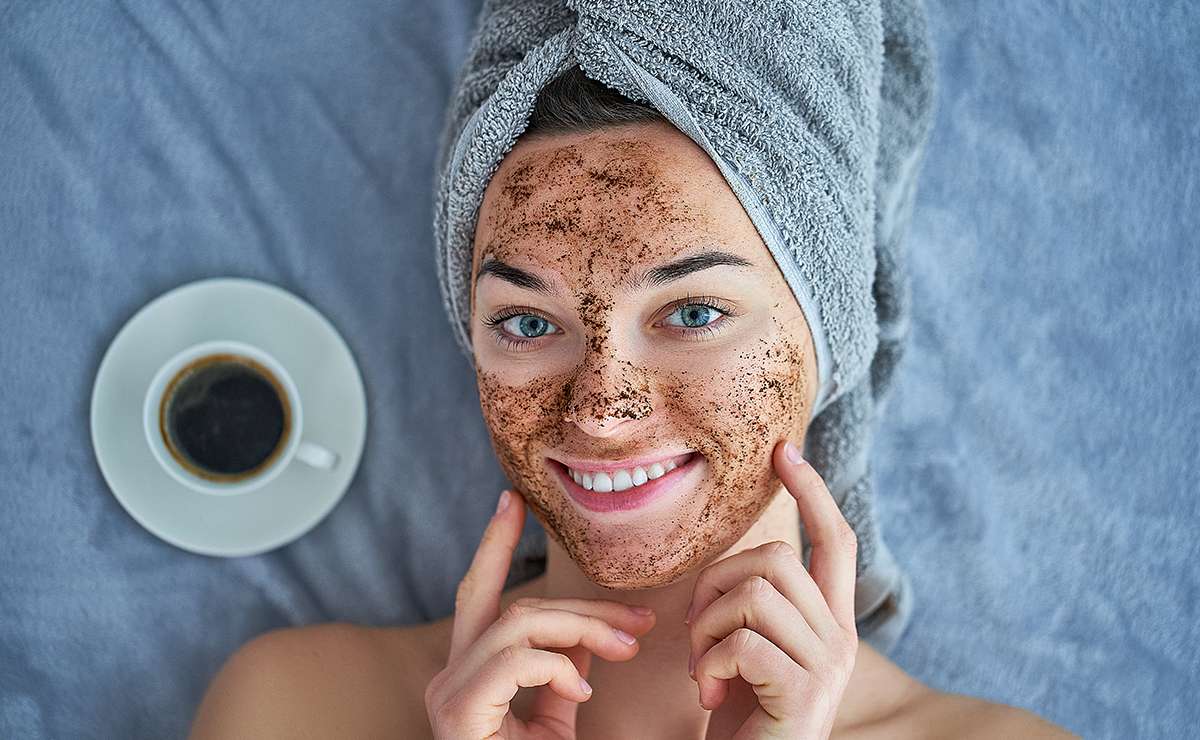 Giovane donna sorridente con un asciugamano avvolto sulla testa e uno scrub naturale fatto con fondi di caffè applicato sul viso, accanto a una tazzina di caffè