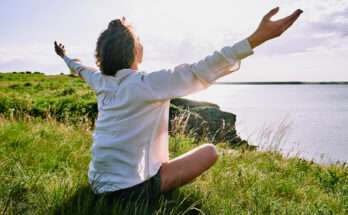 Persona seduta su un prato con le braccia aperte verso il cielo, in atteggiamento di accettazione e benessere, di fronte a un paesaggio naturale