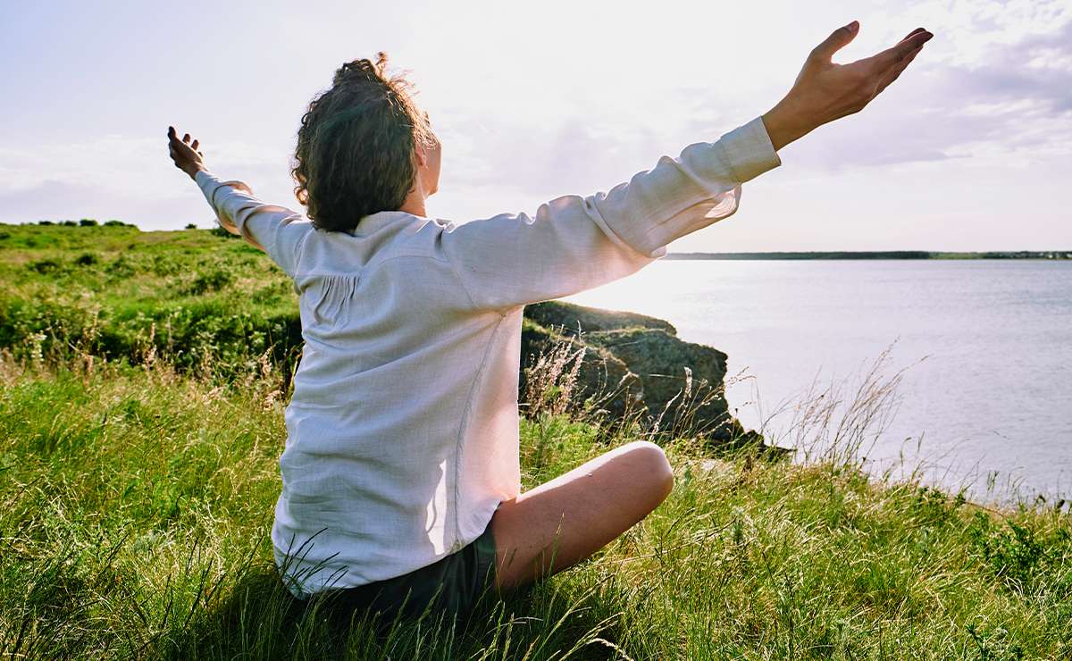 Persona seduta su un prato con le braccia aperte verso il cielo, in atteggiamento di accettazione e benessere, di fronte a un paesaggio naturale