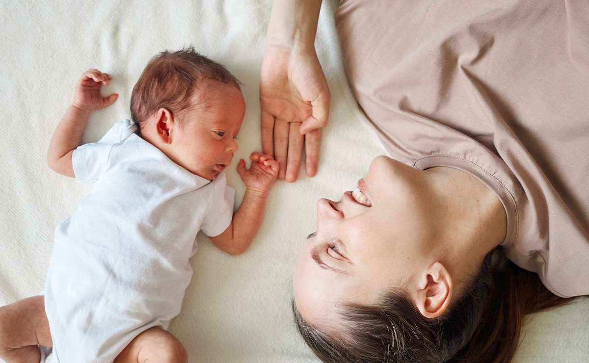 Mamma sdraiata accanto al suo neonato: i due si guardano e si tengono per mano in un gesto tenero e spontaneo che richiama l’istinto materno.