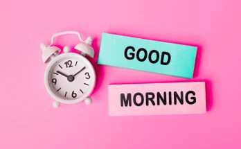 Sveglia bianca con lancette che segnano le 9:10 su sfondo rosa, accanto a due cartoncini colorati con la scritta ‘GOOD MORNING’ in nero.