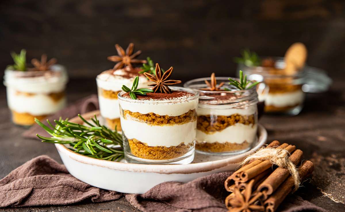 Dolci al cucchiaio serviti in bicchierini di vetro, con strati di crema e biscotti sbriciolati, decorati con anice stellato e rametti di rosmarino. La presentazione è elegante, su un piatto di portata con un tocco rustico di cannella e tessuto marrone.