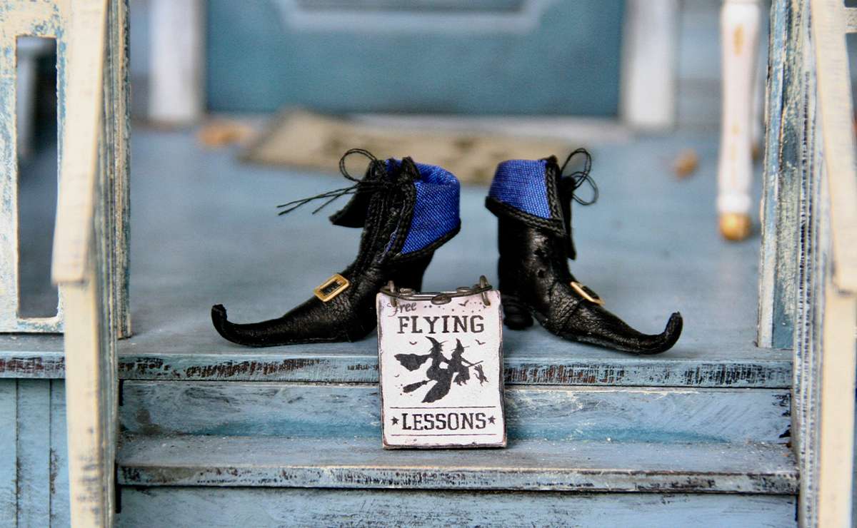 scarpe da Befana nere a punta su un porticato di legno verniciato d'azzurro e davanti una targhetta con raffigurate due befane che volano: la scritta in inglese recita lezioni di volo