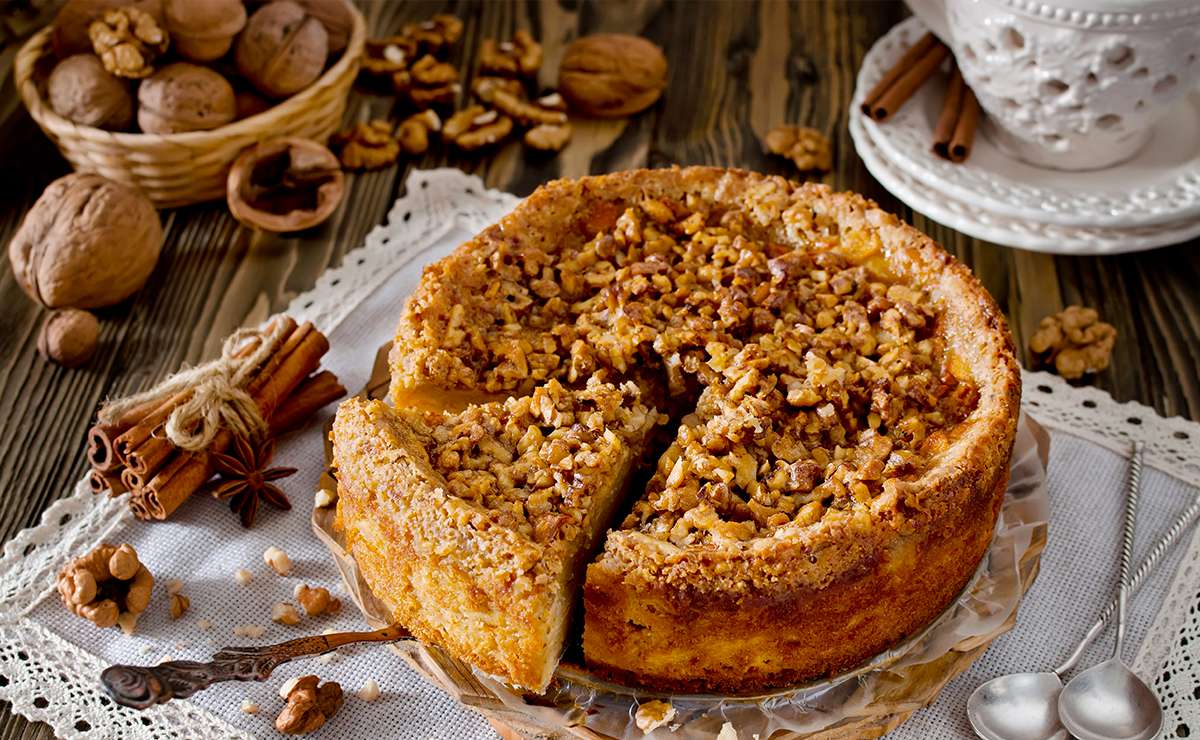 Torta di mele e noci, con una crosta dorata e noci tritate sulla superficie, servita su un piatto con un decorativo tovagliolo di pizzo. Accanto, ci sono noci intere, bastoncini di cannella e un bicchiere decorativo, creando un'atmosfera rustica e accogliente.