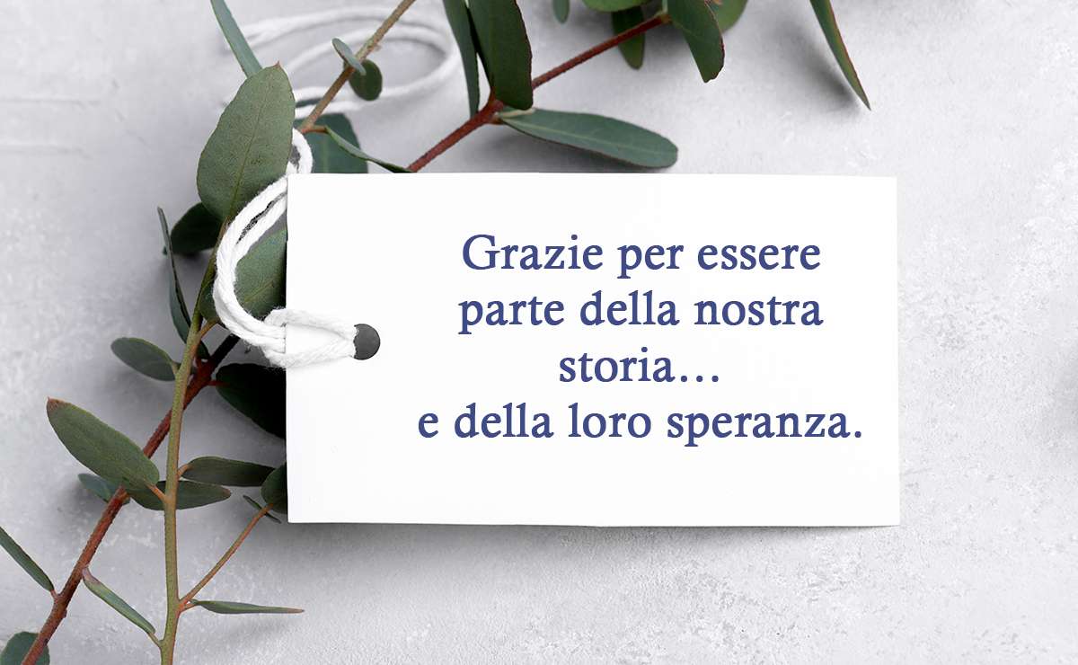 Bigliettino bianco con la frase “Grazie per essere parte della nostra storia… e della loro speranza”, posato su un fondo chiaro decorato con rami verdi.
