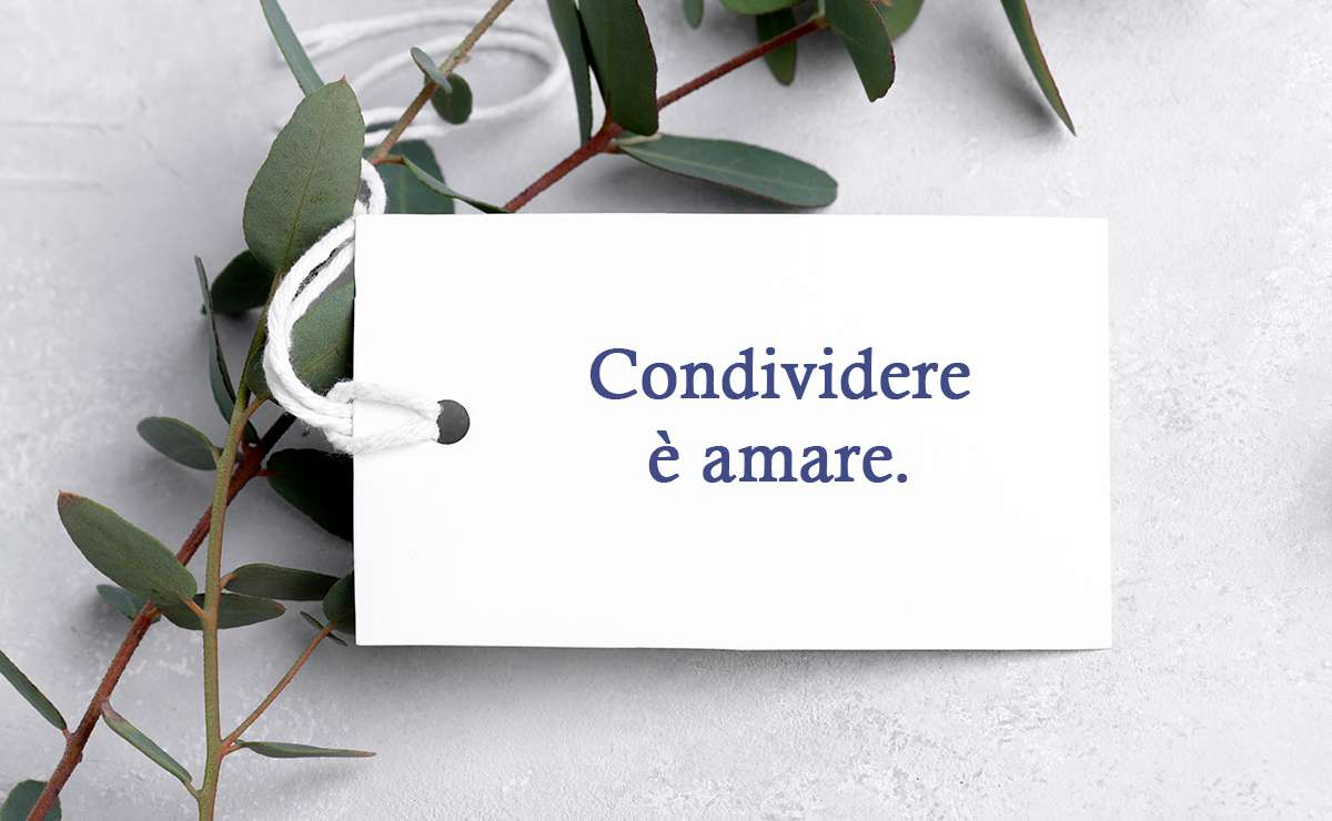 Bigliettino bianco con la frase “Condividere è amare”, posato su un fondo chiaro decorato con rami verdi.