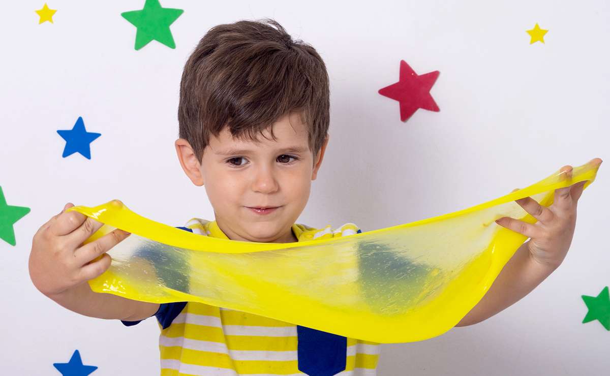 Bambino che gioca con uno slime giallo trasparente, sorridente e concentrato, davanti a uno sfondo decorato con stelle colorate.