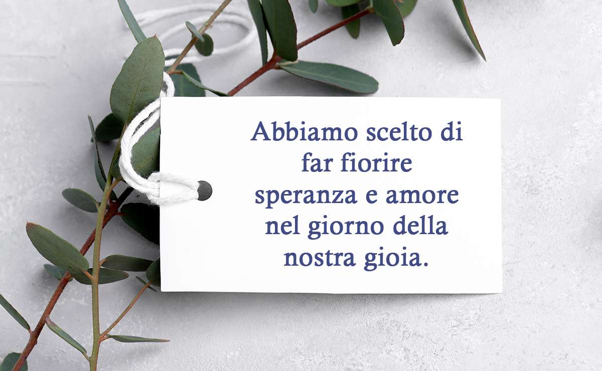 Bigliettino bianco con la frase “Abbiamo scelto di far fiorire speranza e amore nel giorno della nostra gioia”, posato su un fondo chiaro decorato con rami verdi.