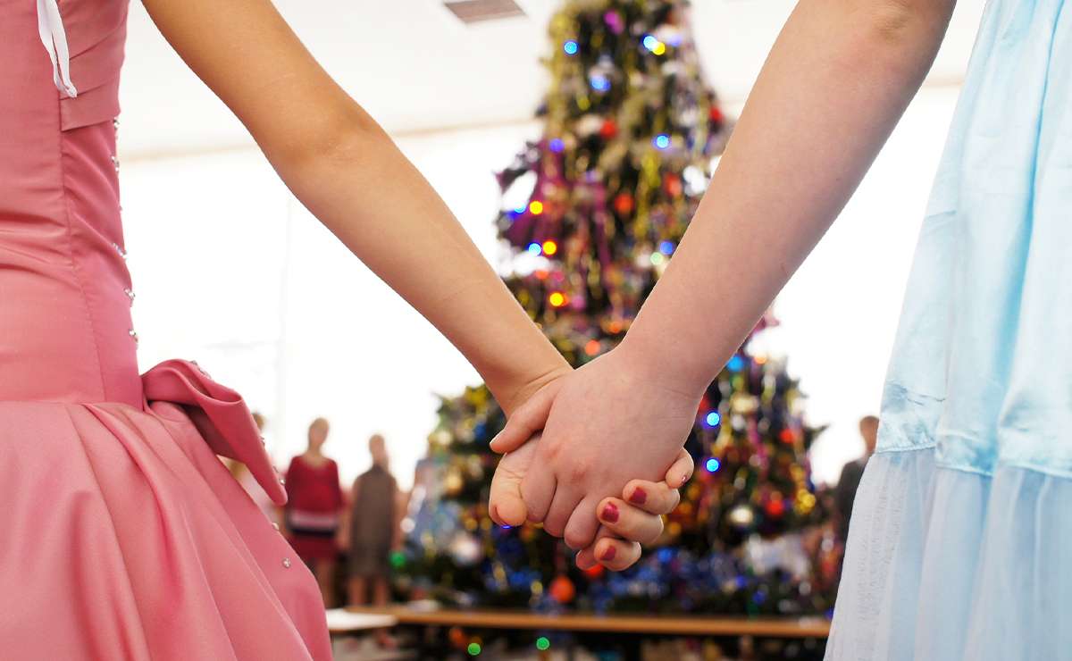 Due bambine che si tengono per mano in primo piano, indossando vestiti eleganti, con un albero di Natale illuminato sullo sfondo.