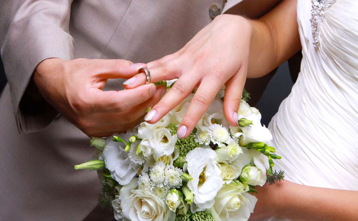Primo piano delle mani di una coppia durante lo scambio delle fedi nuziali: la sposa indossa un abito bianco con dettagli gioiello, mentre lo sposo in abito chiaro infila delicatamente l'anello sull'anulare sinistro della sposa. In primo piano, un bouquet di fiori bianchi composto da rose, lisianthus e fiori di campo verdi.