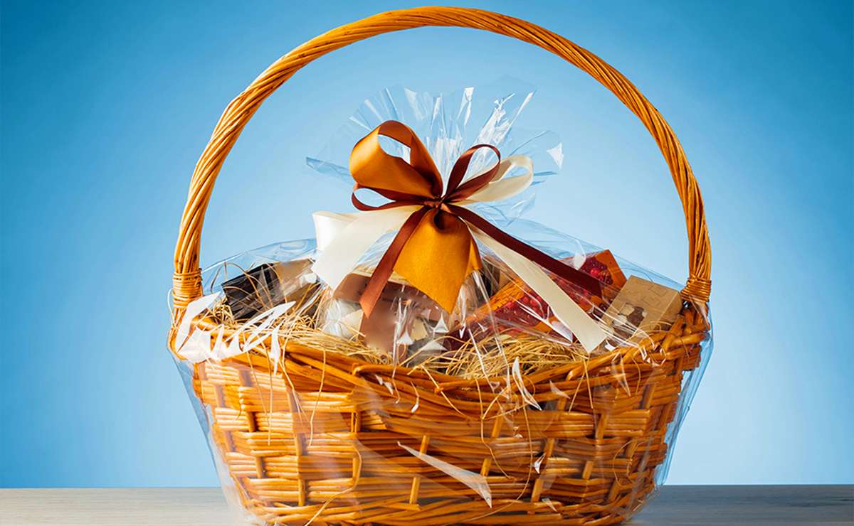 Cesto regalo gastronomico natalizio in vimini avvolto in cellophane trasparente, decorato con un fiocco marrone e crema, contenente prodotti gastronomici su sfondo azzurro.