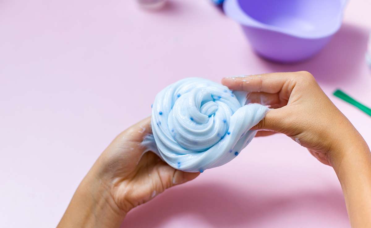 Mani che modellano uno skifidol slime morbido e azzurro con piccole sfere blu, su uno sfondo rosa pastello, con ciotole e strumenti sullo sfondo.