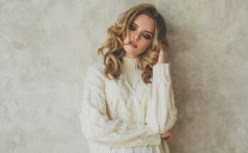 Donna con balayage capelli biondi e morbidi ricci, indossa un maglione bianco a trecce e posa con espressione serena davanti a un muro chiaro con texture.