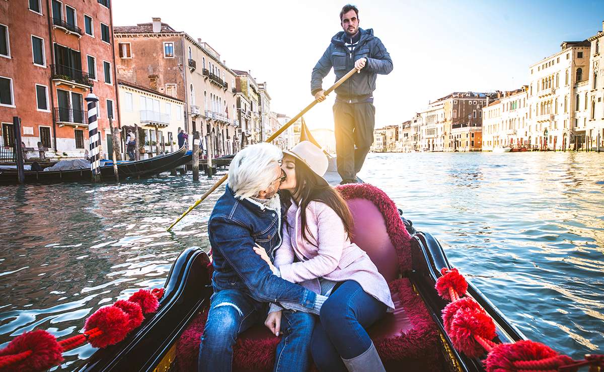 Una coppia innamorata si bacia teneramente a bordo di una gondola adornata di velluto rosso, mentre un gondoliere rema sul Canal Grande a Venezia. Sullo sfondo si intravedono gli edifici storici della città e l’acqua scintillante al sole, creando un’atmosfera romantica perfetta per San Valentino in una delle mete più iconiche d’Italia.