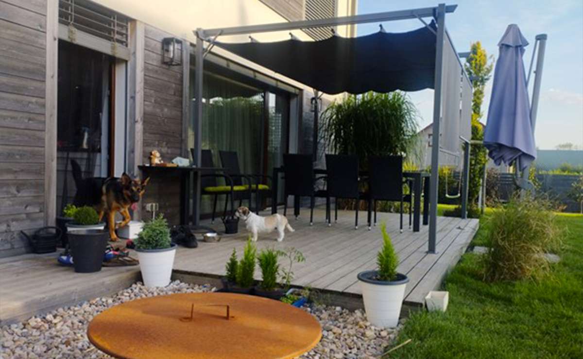 Terrazza in legno con pergola moderna, tavolo da esterno con sedie nere e cuscini verdi, decorata con piante in vaso. Due cani passeggiano nello spazio verde circostante.