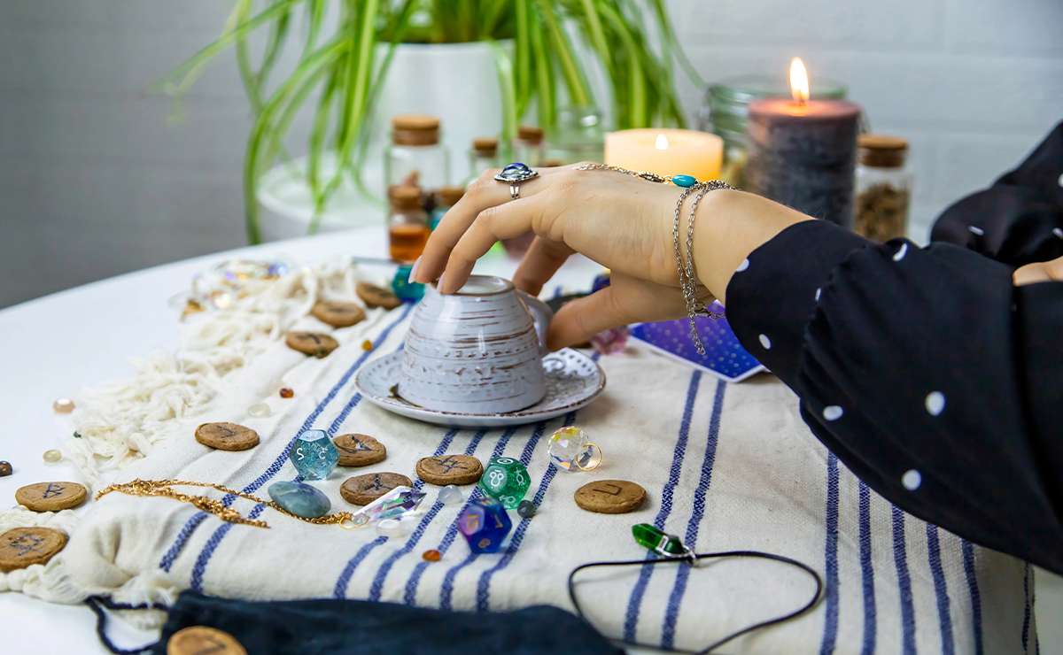 Mano femminile solleva una tazzina rovesciata su un piattino durante una lettura di caffèomanzia, l'arte della lettura dei fondi di caffè, con decorazioni esoteriche, cristalli e candele accese su una tovaglia a righe