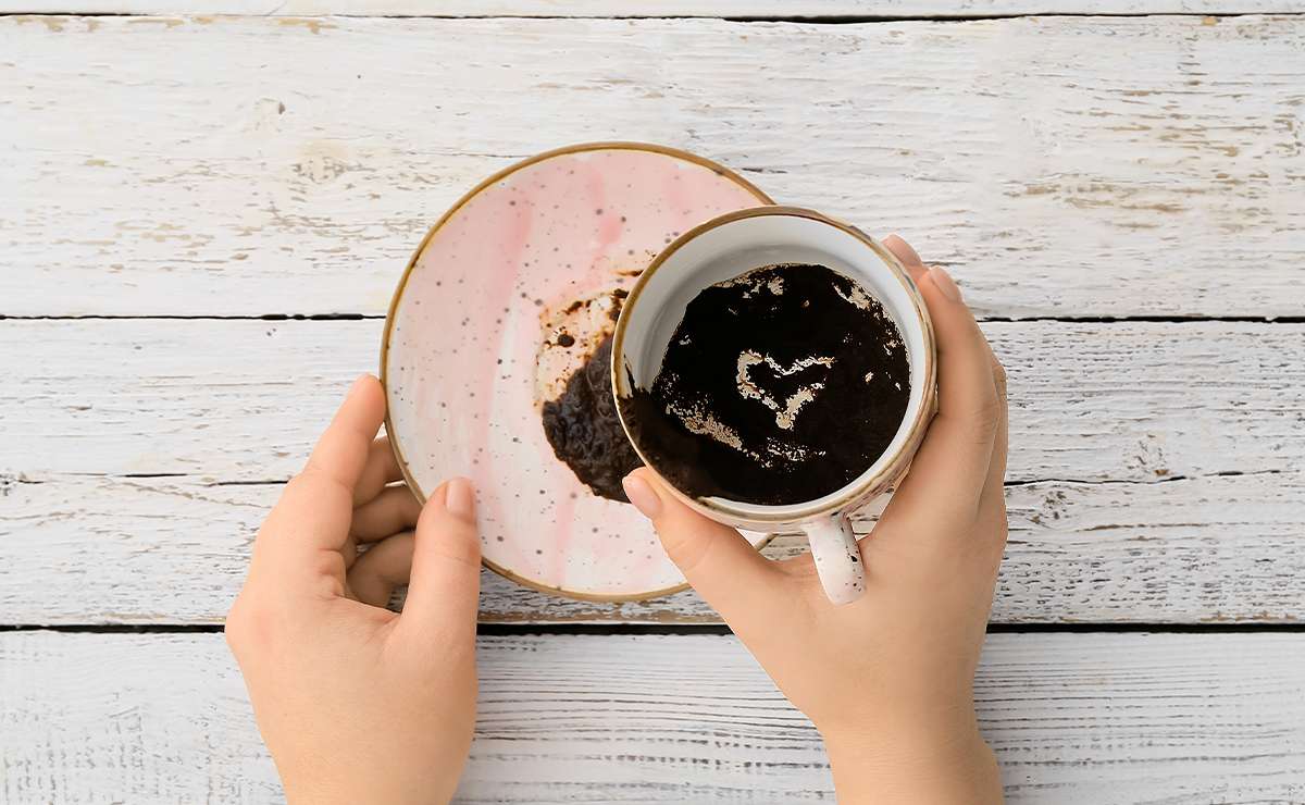 Mani che tengono una tazza di caffè con fondi visibili e un piattino sottostante, durante una lettura simbolica dei fondi di caffè su un tavolo in legno chiaro.