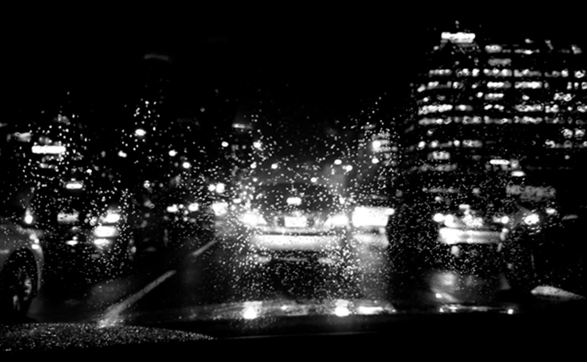 Vista di una strada trafficata di notte sotto la pioggia, con gocce d'acqua sul parabrezza e luci sfocate delle auto e degli edifici.