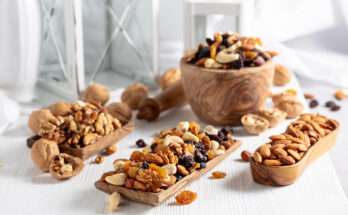 Mix di frutta secca su vassoi di legno, con mandorle, noci, anacardi e uvetta, disposti su un tavolo bianco accanto a ciotole in legno e decorazioni.