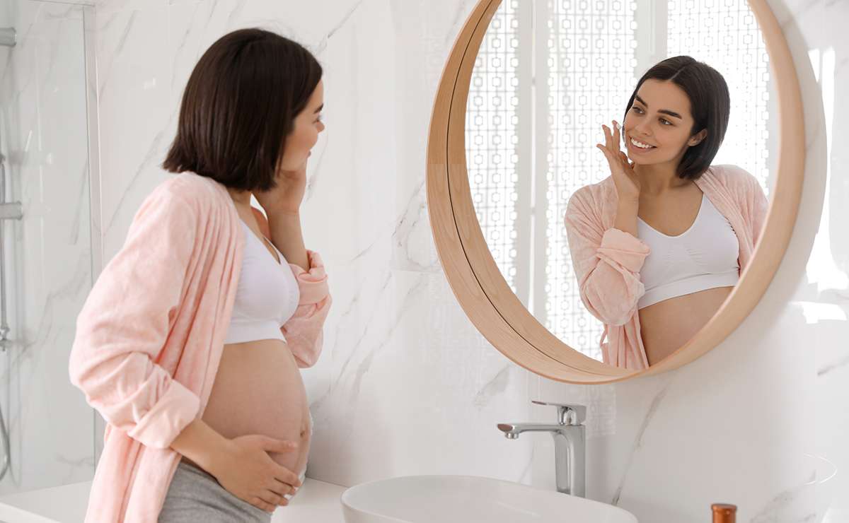 Donna incinta che si guarda allo specchio e accarezza il viso, riflettendo sulla cura della pelle durante la gravidanza in un bagno luminoso.