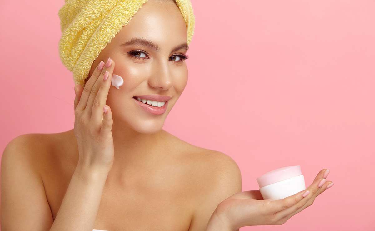 Donna con asciugamano giallo sui capelli applica crema viso davanti a sfondo rosa, tenendo in mano un barattolo di crema bianca con coperchio rosa.