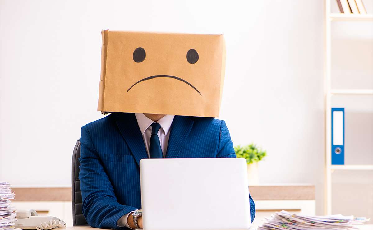 Uomo in abito elegante seduto alla scrivania con un computer portatile, indossa una scatola di cartone sulla testa con una faccia triste disegnata, simbolo della sindrome dell'impostore e dell'insicurezza lavorativa.