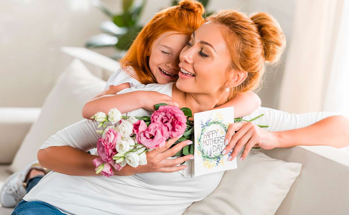 Bambina dai capelli rossi abbraccia la sua mamma sorridente sul divano per la Festa della Mamma. La madre tiene in mano un biglietto con la scritta ‘Happy Mother’s Day’ e un mazzo di fiori rosa e bianchi.