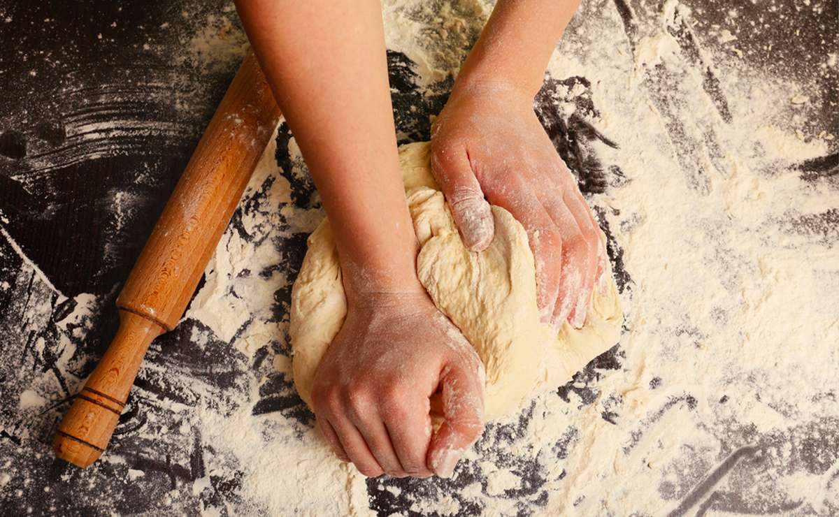 Mani che impastano la pasta su una superficie infarinata, con un mattarello di legno accanto. Scena tradizionale di preparazione artigianale della pasta.