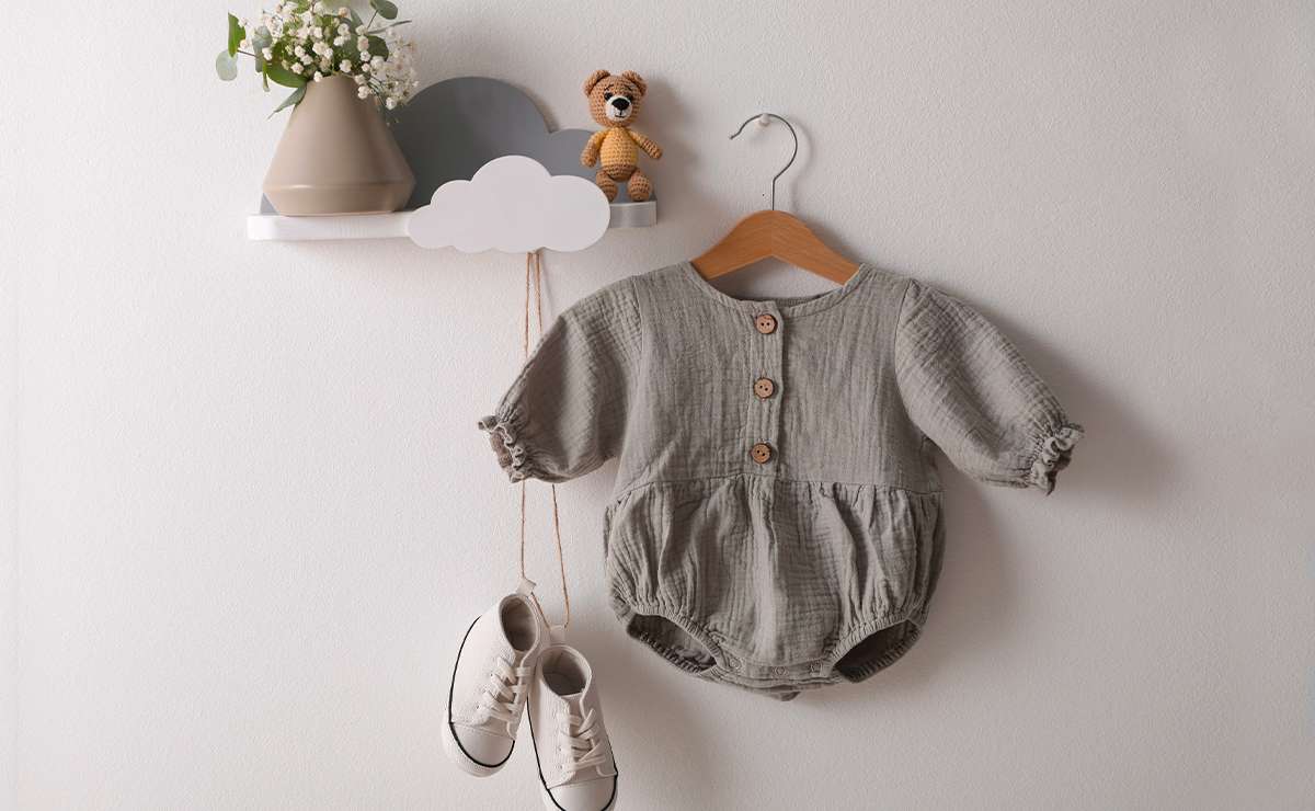 Pagliaccetto estivo per neonato in mussola di cotone grigio-verde con bottoni in legno e maniche arricciate, appeso a una gruccia su sfondo bianco, accanto a un paio di scarpine bianche e una mensola con decorazioni baby.