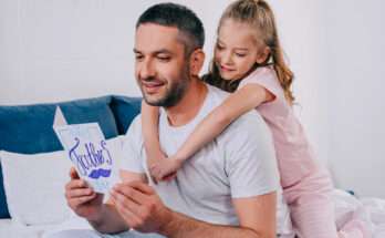 Padre sorridente legge un biglietto con la scritta “Happy Father’s Day” mentre la figlia lo abbraccia da dietro con affetto.