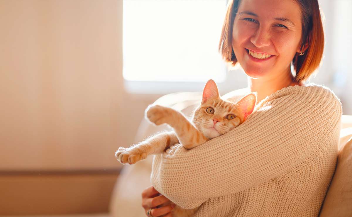 Donna sorridente abbraccia un gatto arancione in una stanza illuminata dal sole, simbolo di affetto e complicità tra umano e felino per la Festa del Gatto