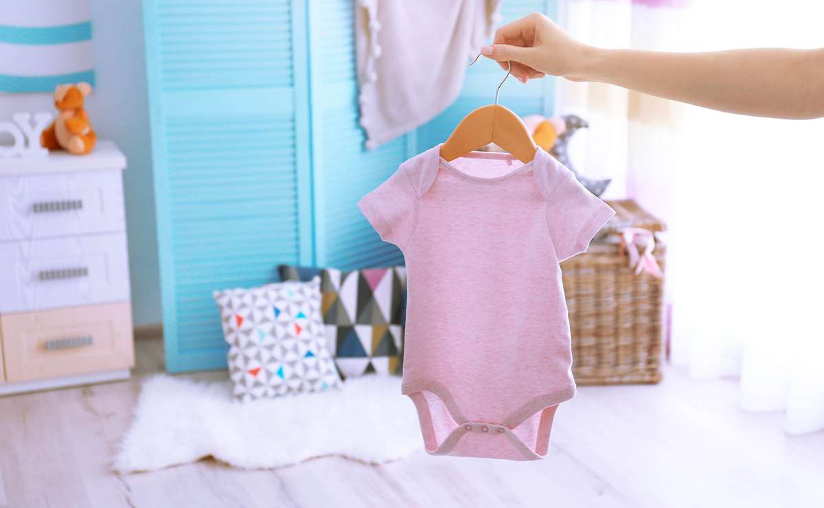 Mano che regge una gruccia con un body rosa per neonato, in una cameretta luminosa con arredamento azzurro e decorazioni colorate.