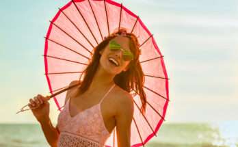 Donna sorridente con occhiali a forma di cuore e ombrellino rosa sulla spiaggia al tramonto, simbolo di spensieratezza estiva.