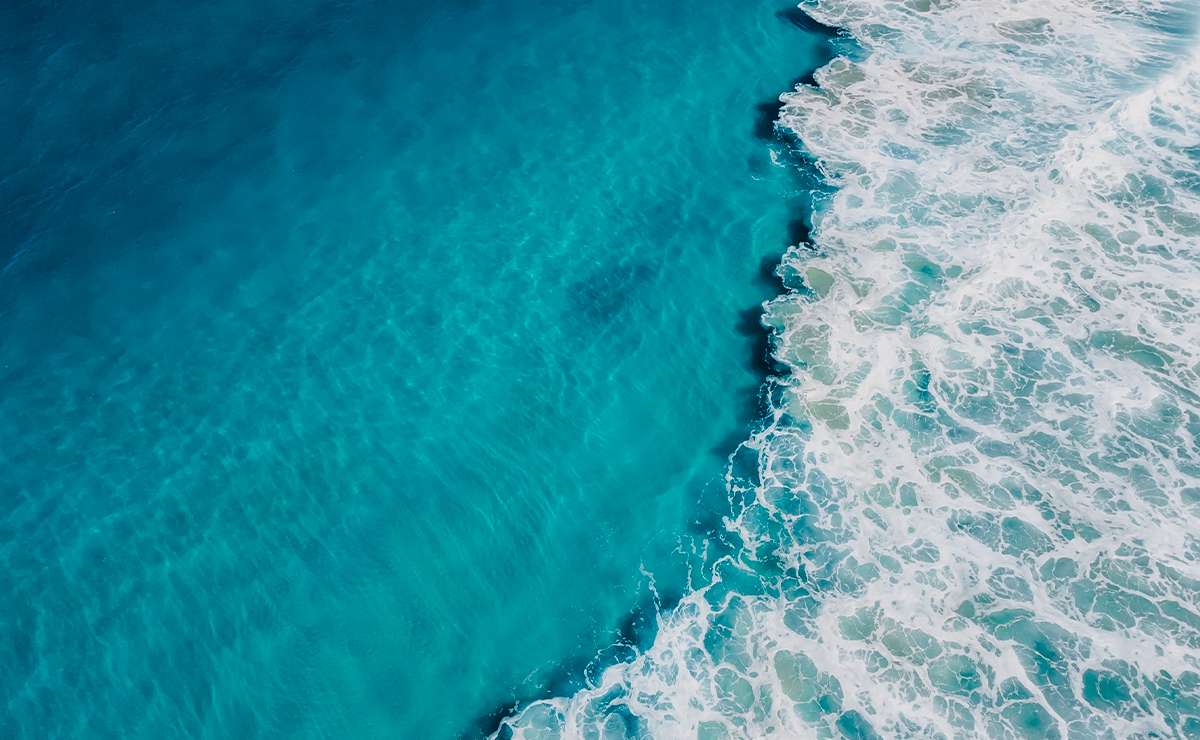 Vista aerea dell’oceano con acqua cristallina e onde bianche che si infrangono, creando un contrasto tra il blu profondo e la schiuma marina.