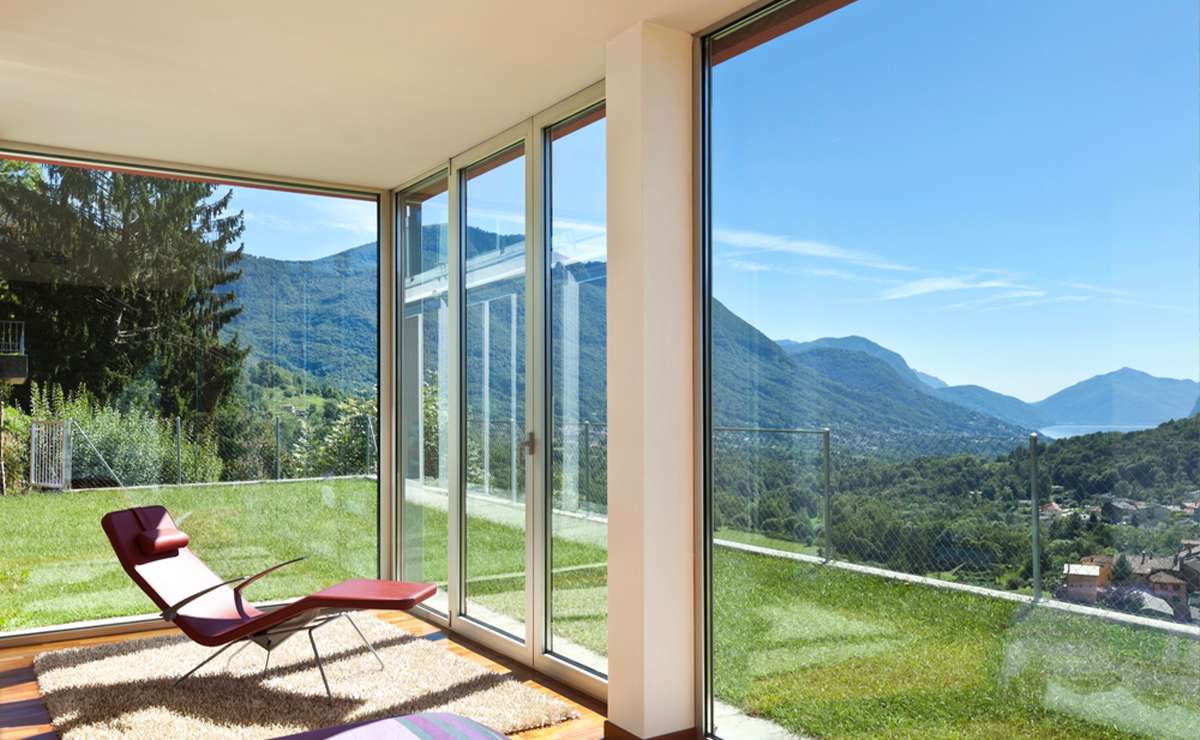 Interno di un giardino d’inverno moderno con ampie vetrate panoramiche, vista su montagne e vallata, poltrona design e tappeto su pavimento in legno.