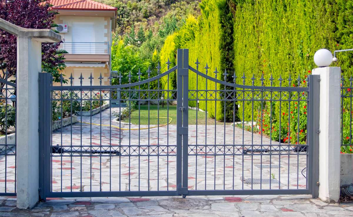 Cancello in ferro battuto moderno all’ingresso di una villetta con vialetto pavimentato e giardino curato.