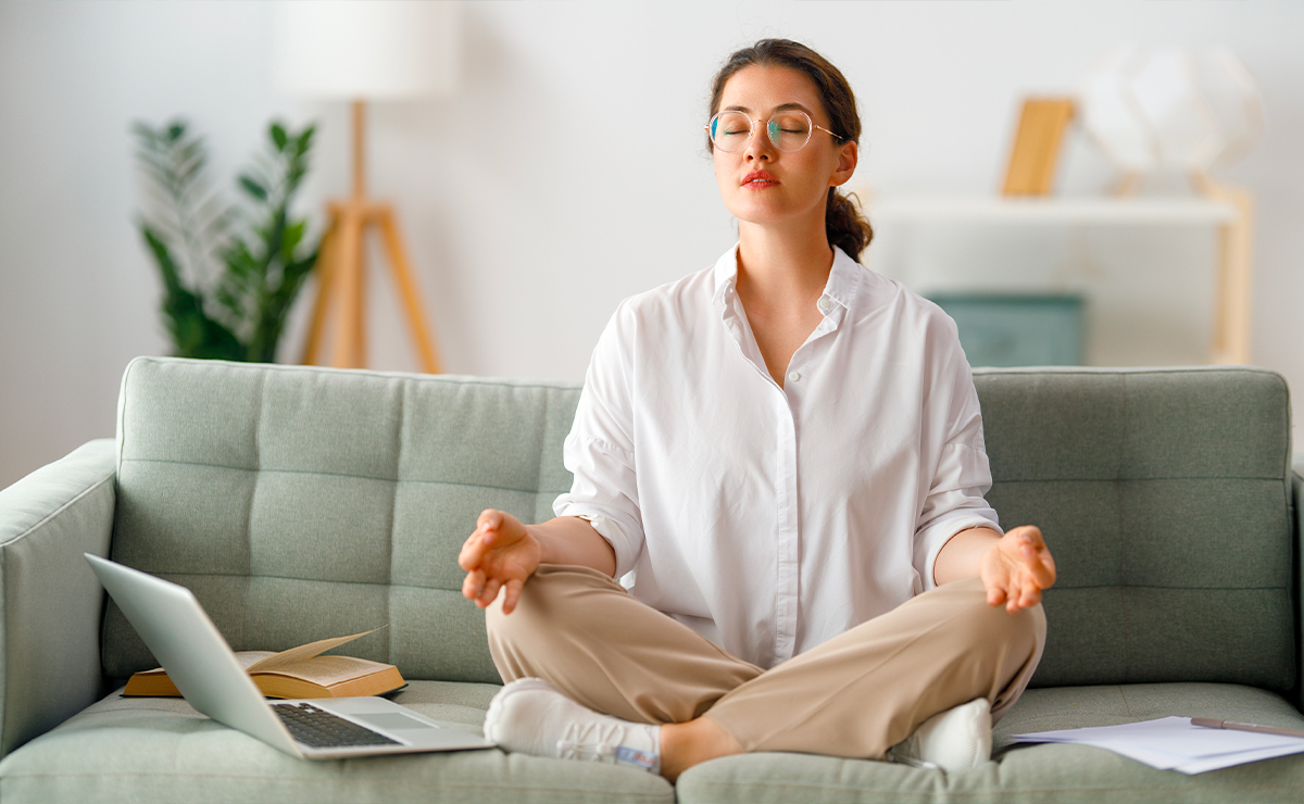 Donna seduta sul divano in posizione meditativa, occhi chiusi e aria serena, mentre pratica un momento di consapevolezza e introspezione, con laptop e libro accanto.