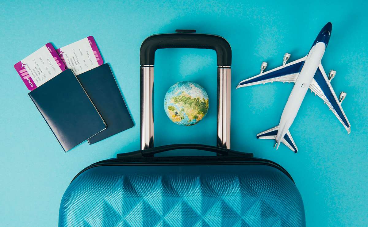 Valigia rigida blu con manico estraibile, due passaporti con carte d’imbarco, un mappamondo e un modellino di aereo su sfondo azzurro.