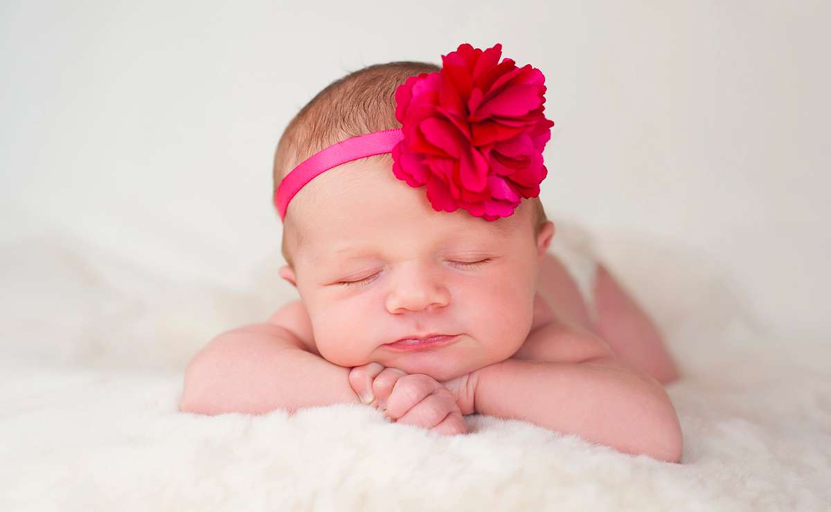 Bambina neonata addormentata con fascia rosa e fiore rosso tra i capelli, sdraiata su una coperta morbida bianca.