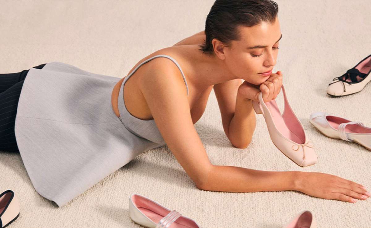 Modella sdraiata su tappeto chiaro mentre osserva una ballerina rosa, circondata da altre calzature flat, tendenza moda primavera estate 2025.
