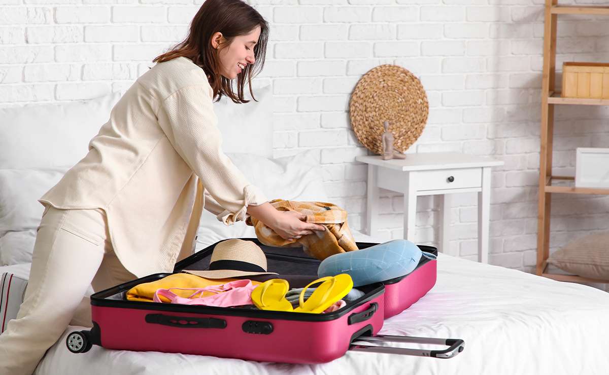 Donna che prepara la valigia su un letto, inserendo vestiti estivi e accessori da viaggio in un trolley rosa, in una stanza luminosa e ordinata con pareti bianche.